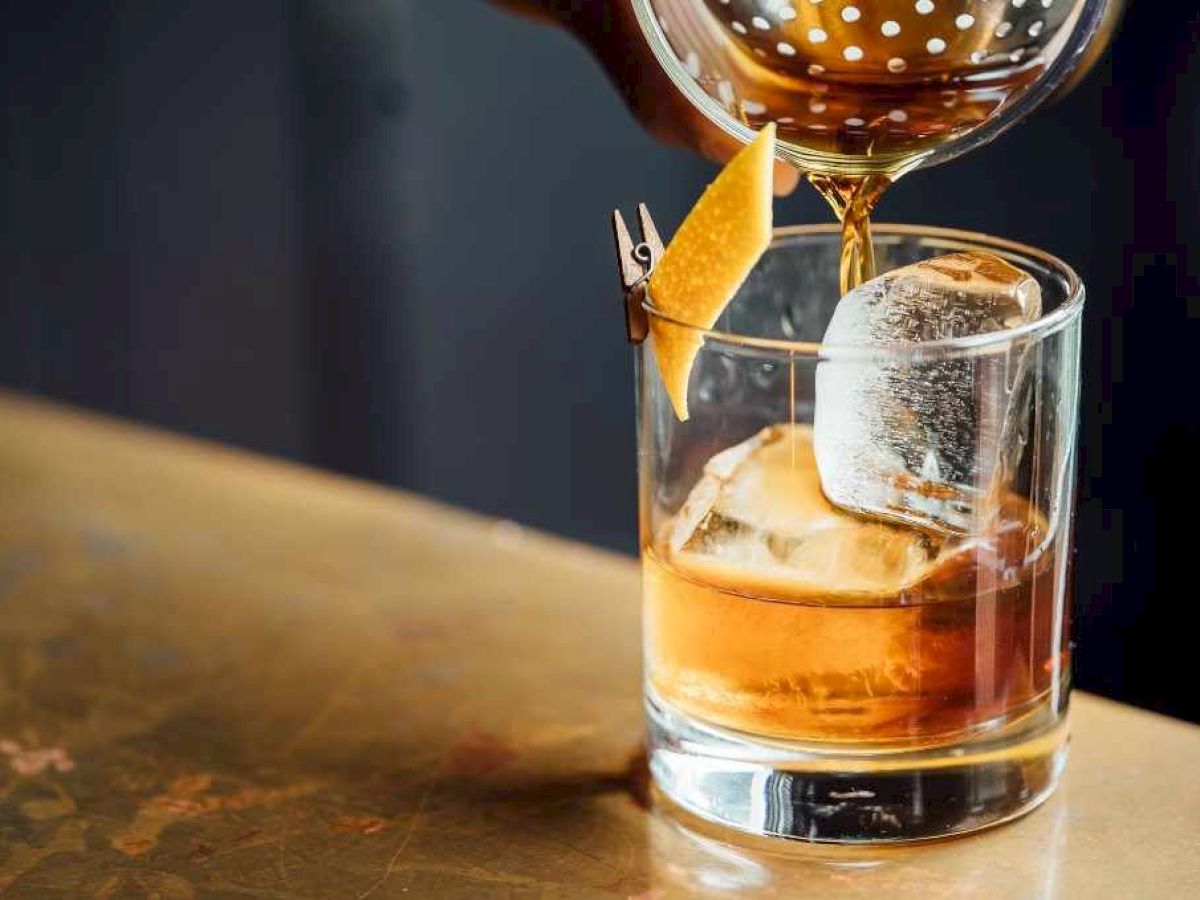 A cocktail is being poured into a glass with ice and garnished with a lemon twist, set on a table.