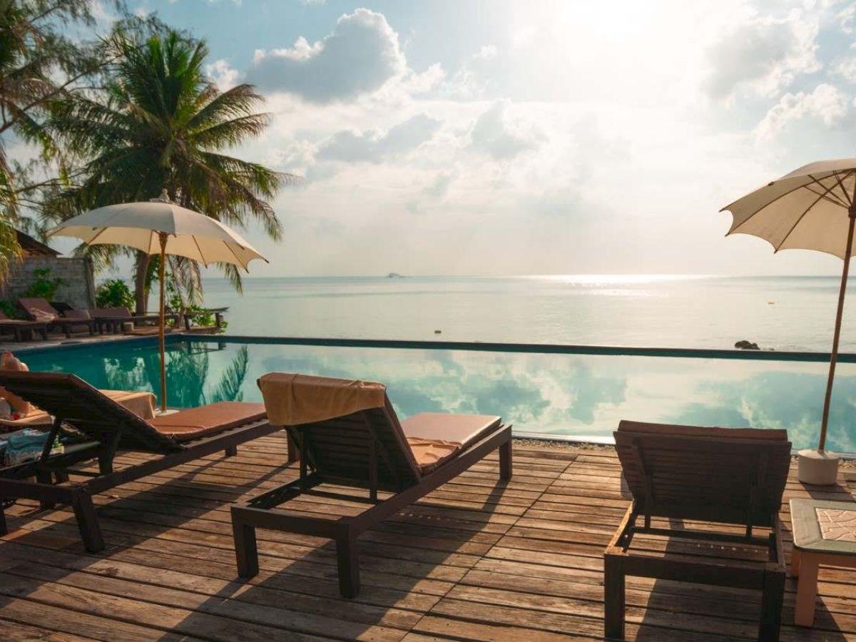 The image shows a serene beach scene with sun loungers, umbrellas, a pool, and palm trees overlooking a calm ocean under a partly cloudy sky.