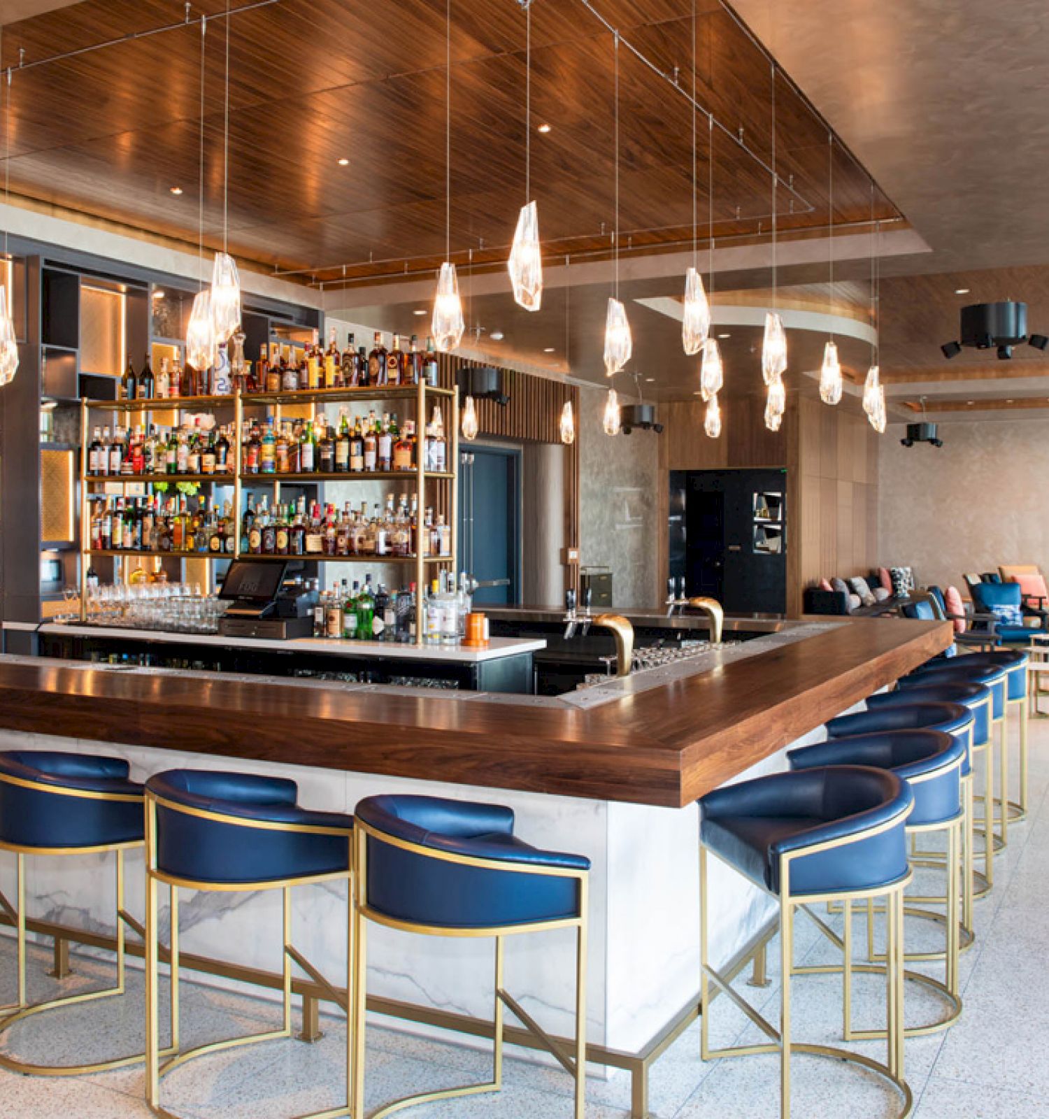 A stylish bar with a wooden counter, blue chairs, hanging lights, and a well-stocked wall of bottles in a modern, open space.