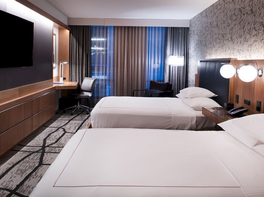 The image shows a modern hotel room with two beds, a wall-mounted TV, and a work desk by the window, featuring stylish decor.