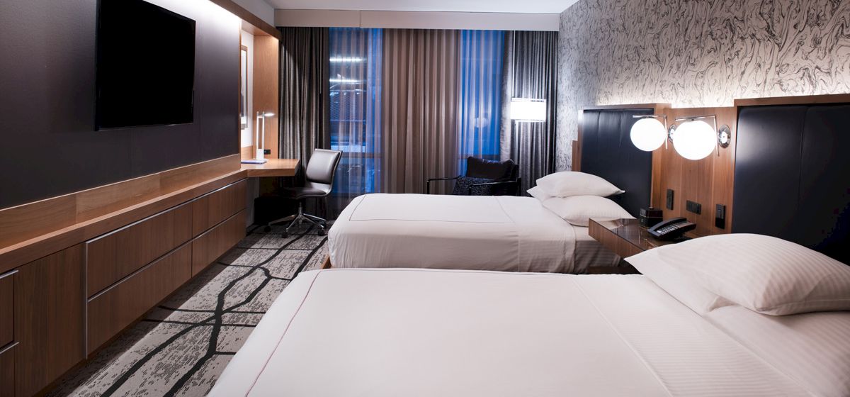 The image shows a modern hotel room with two beds, a wall-mounted TV, and a work desk by the window, featuring stylish decor.