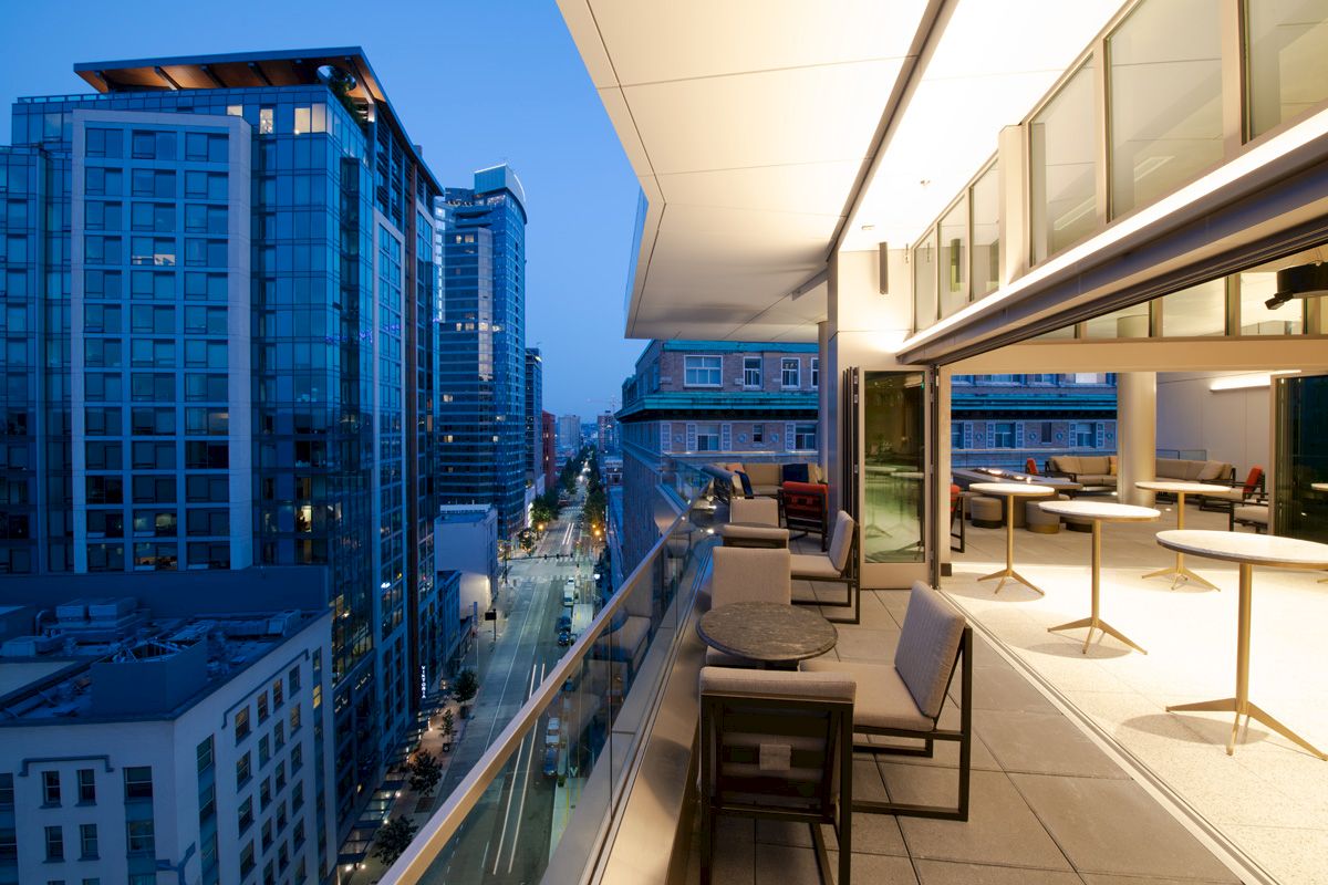 A modern balcony with seating overlooks a city street lined with tall buildings at dusk, offering a stylish urban view.