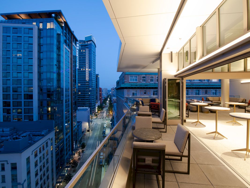 A modern balcony with seating overlooks a city street lined with tall buildings at dusk, offering a stylish urban view.