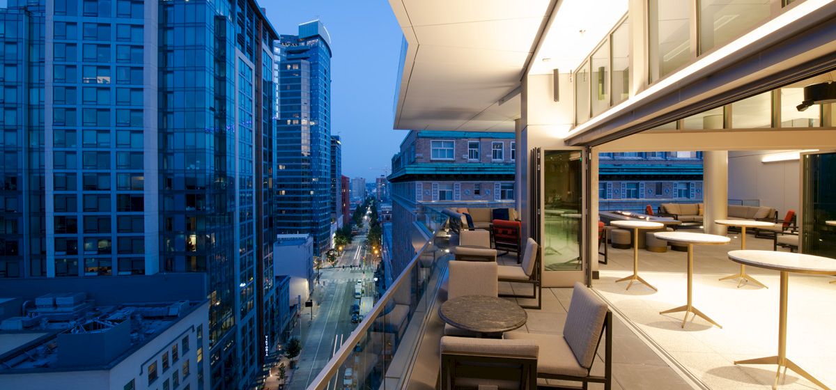 A modern balcony with seating overlooks a city street lined with tall buildings at dusk, offering a stylish urban view.