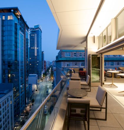A modern balcony view of a cityscape at dusk, featuring outdoor seating, buildings, and a lit walkway.