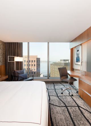 A modern hotel room with a large bed, wall-mounted TV, desk, and a window view of city buildings and the water.