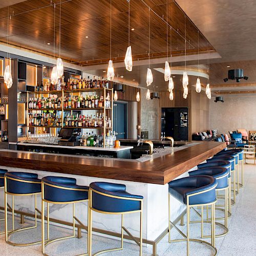 A modern bar with a wooden counter, blue stools, hanging lights, and a well-stocked shelf of bottles, in a spacious, stylish room.