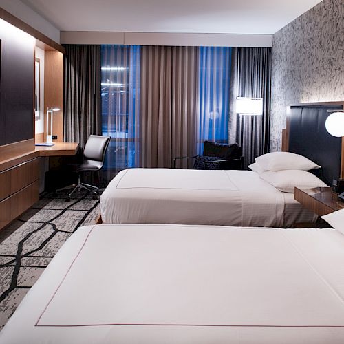 The image shows a hotel room with two beds, a desk, a lamp, curtains, a TV on the wall, and patterned wallpaper.