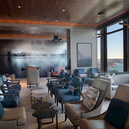 A modern lounge with leather armchairs, a cityscape mural, and large windows offering a view of a body of water and a Ferris wheel.