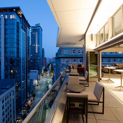 Rooftop patio overlooking a cityscape with tall buildings, featuring seating and tables, captured at dusk with lights illuminating the scene.