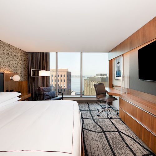 A modern hotel room features a large bed, TV, desk, and a window with a cityscape view, showcasing a sleek and stylish interior design.