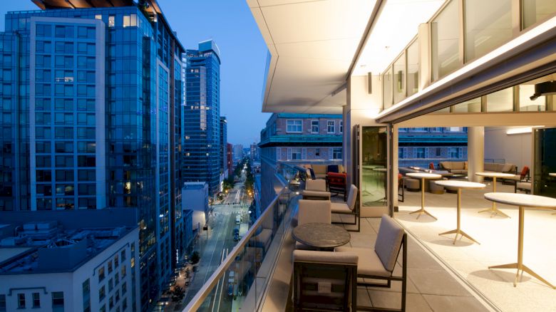 A modern cityscape at dusk with tall buildings, a lit balcony featuring seating and tables, and a view of the street below.