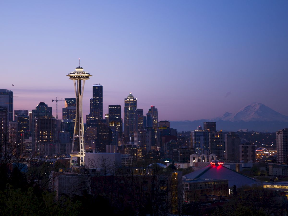 A city skyline at dusk featuring a prominent tower, surrounded by skyscrapers, with a mountain in the background under a twilight sky.