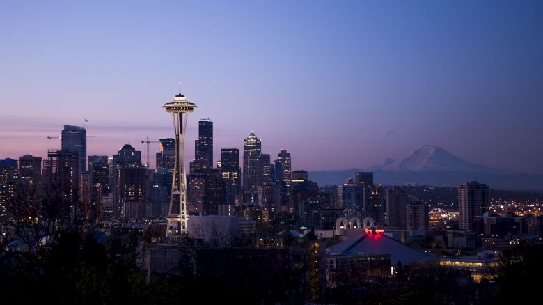A city skyline at dusk featuring a prominent tower, surrounded by skyscrapers, with a mountain in the background under a twilight sky.