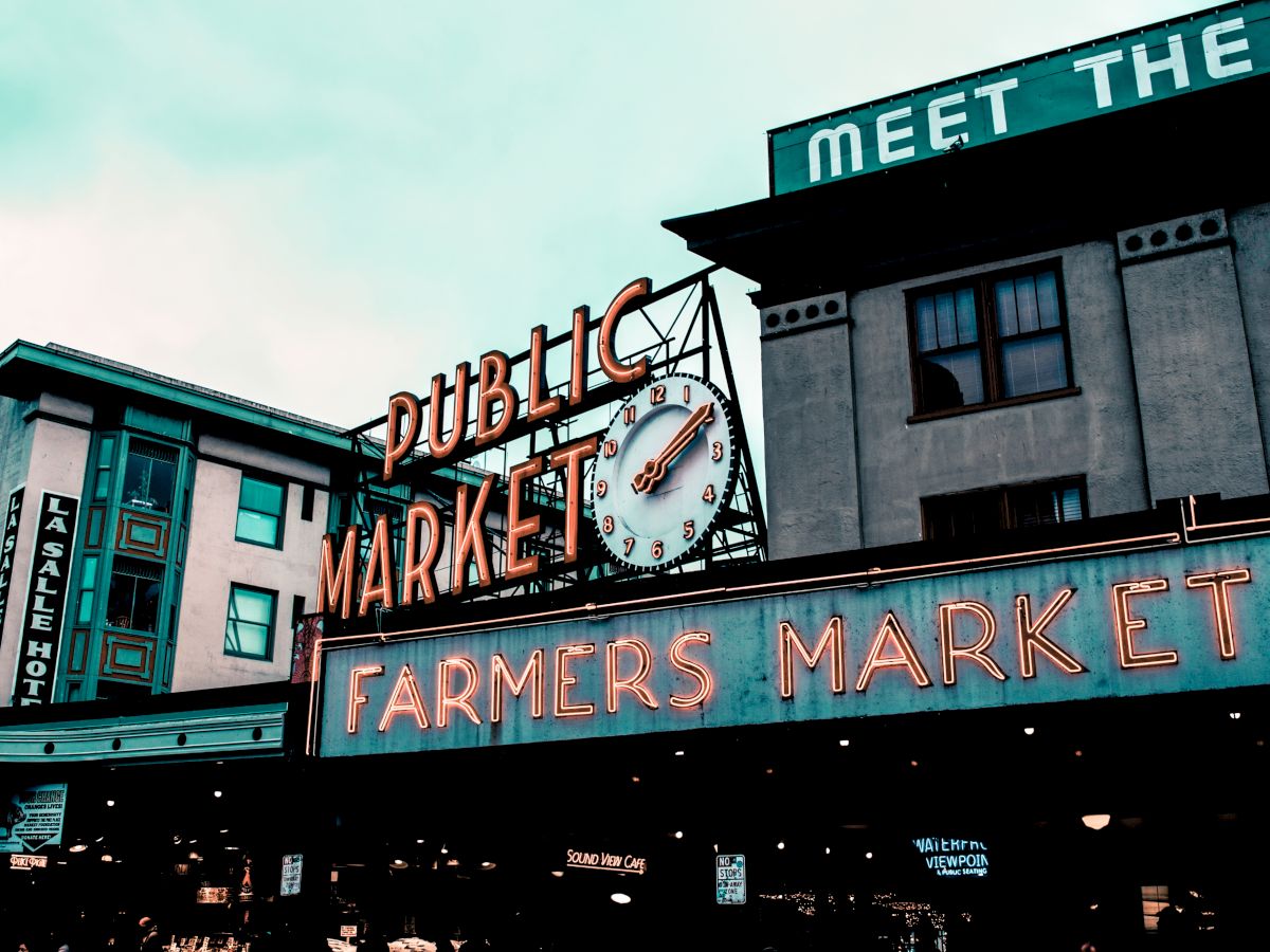 The image shows an exterior view of a building with signs for a "Public Market" and "Farmers Market" displayed prominently in neon lights.