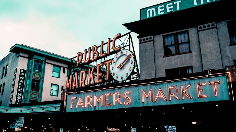 The image shows an exterior view of a building with signs for a "Public Market" and "Farmers Market" displayed prominently in neon lights.