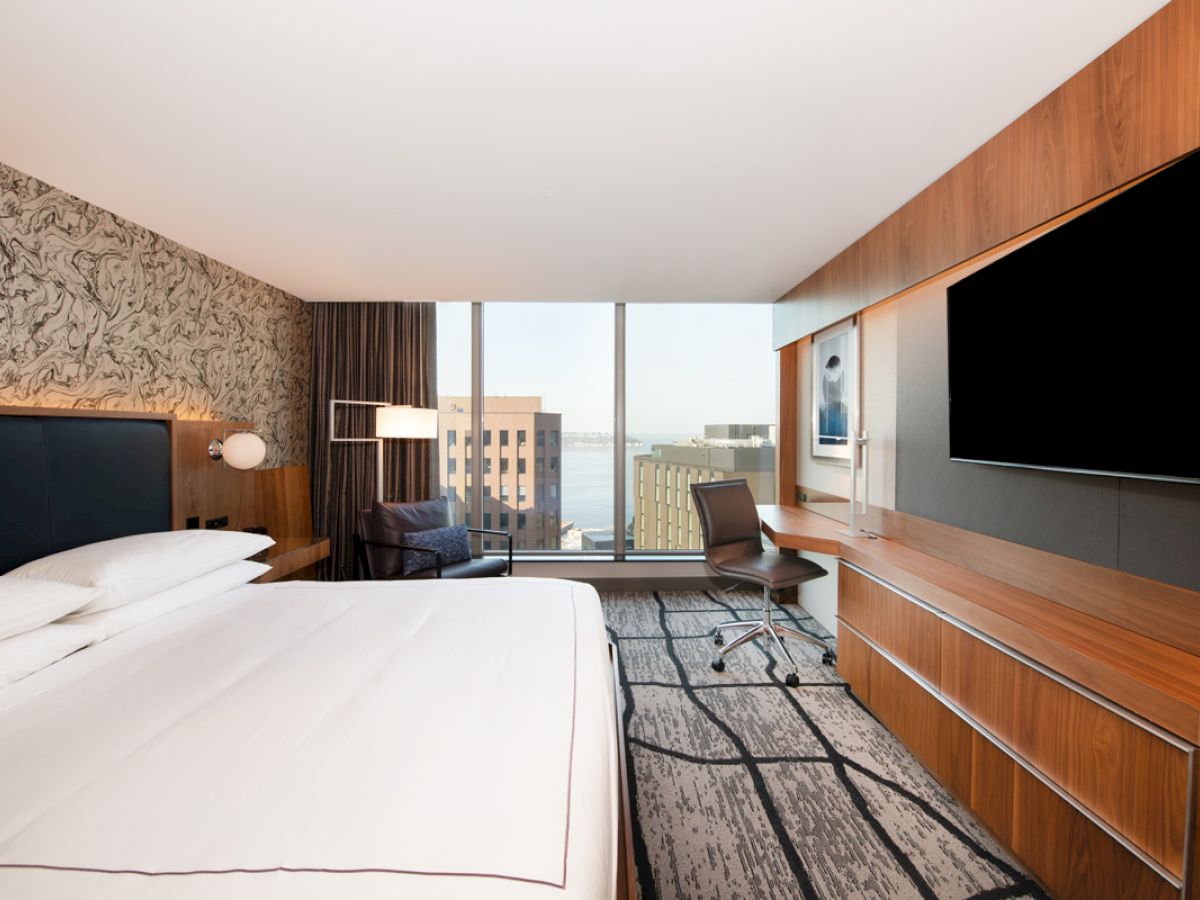 A modern hotel room features a large bed, stylish decor, a desk with a chair, and a big window with a view of buildings.