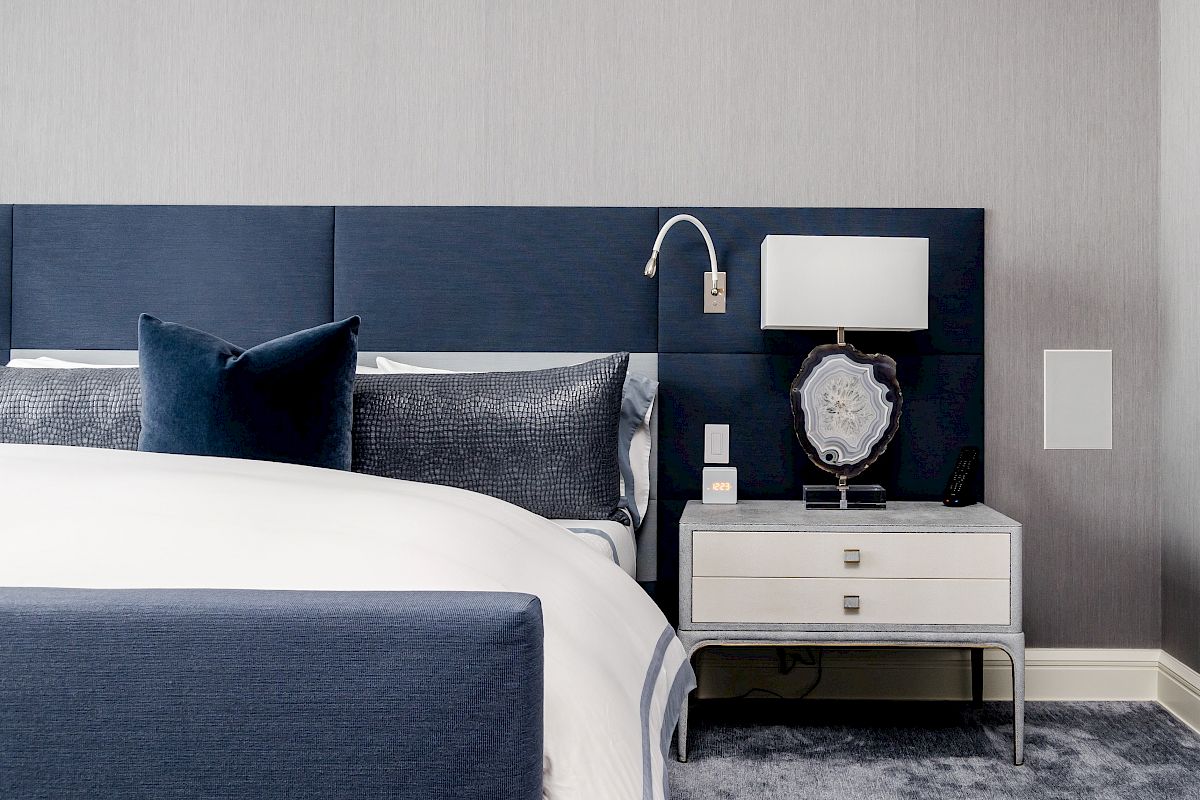 A modern bedroom with a navy blue headboard, white bedding, and a nightstand with a lamp and decorative items sits on a plush carpet.
