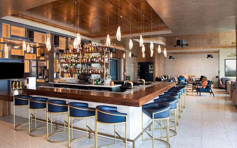 A stylish bar with blue chairs, pendant lights, and a stocked back shelf in a modern room with seating areas and sleek decor.