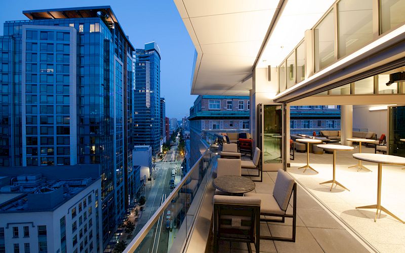 A city skyline view from a modern balcony with seating and tables, adjacent to tall buildings during twilight.