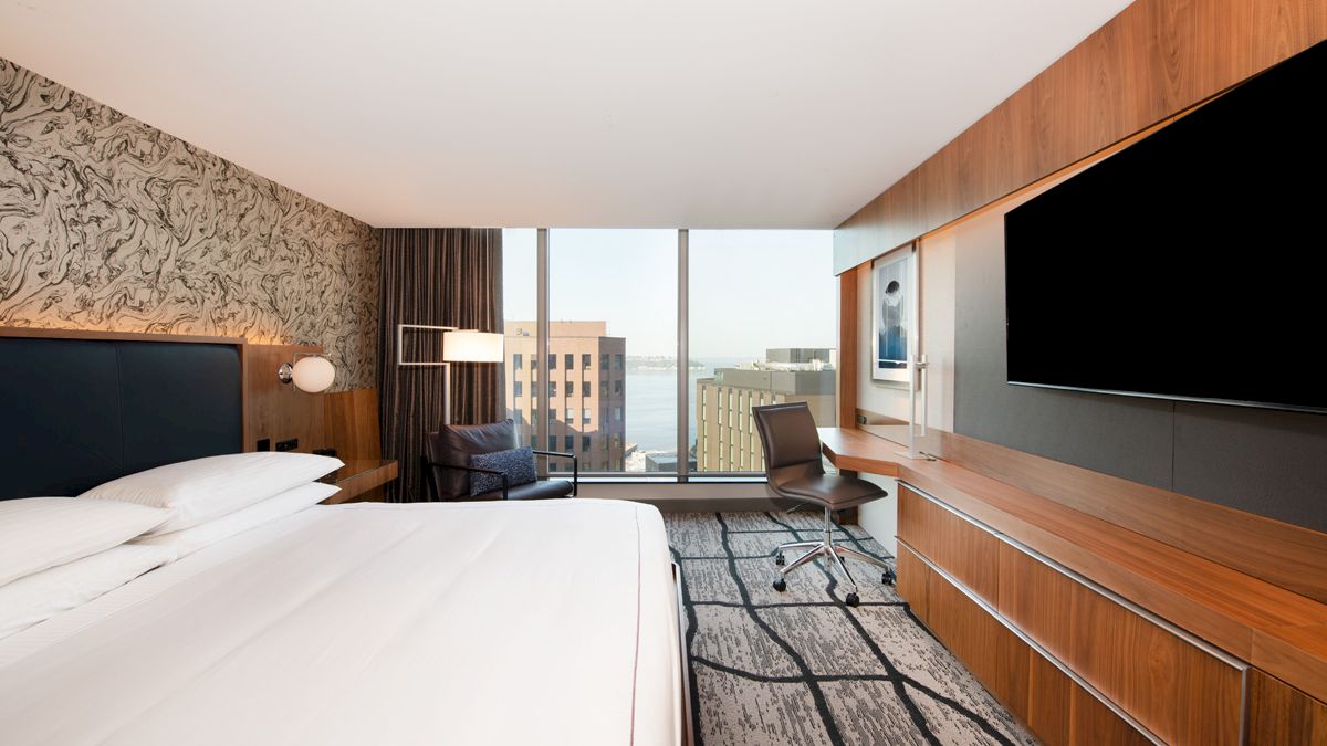A modern hotel room with a bed, desk, chair, large TV, and window view of buildings and water, featuring stylish decor and wood accents.