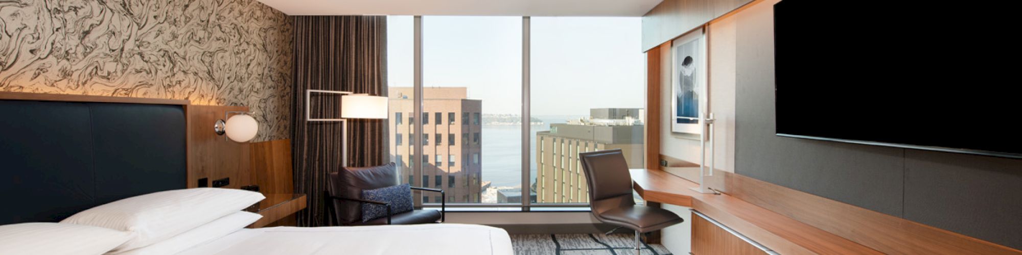 A modern hotel room with a bed, desk, chair, large TV, and window view of buildings and water, featuring stylish decor and wood accents.